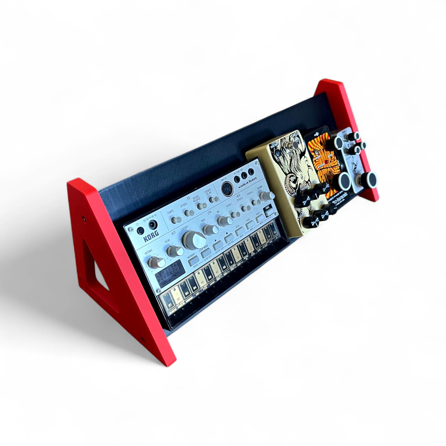 3D printed Desktop Pedalboard 400mm x 150mm 60 Degree Guitar Pedal & Synth Stand stand by Cold Earth Industries, shown in black with red sides on a white background. The stand is holding a Korg Volca Bass and guitar pedals – custom desktop stands.