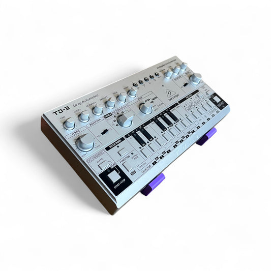 3D printed Behringer TD-3 / RD-6 / RD-78 Synth 30 degree stand by Cold Earth Industries, shown in purple on a white background holding a TD-3
