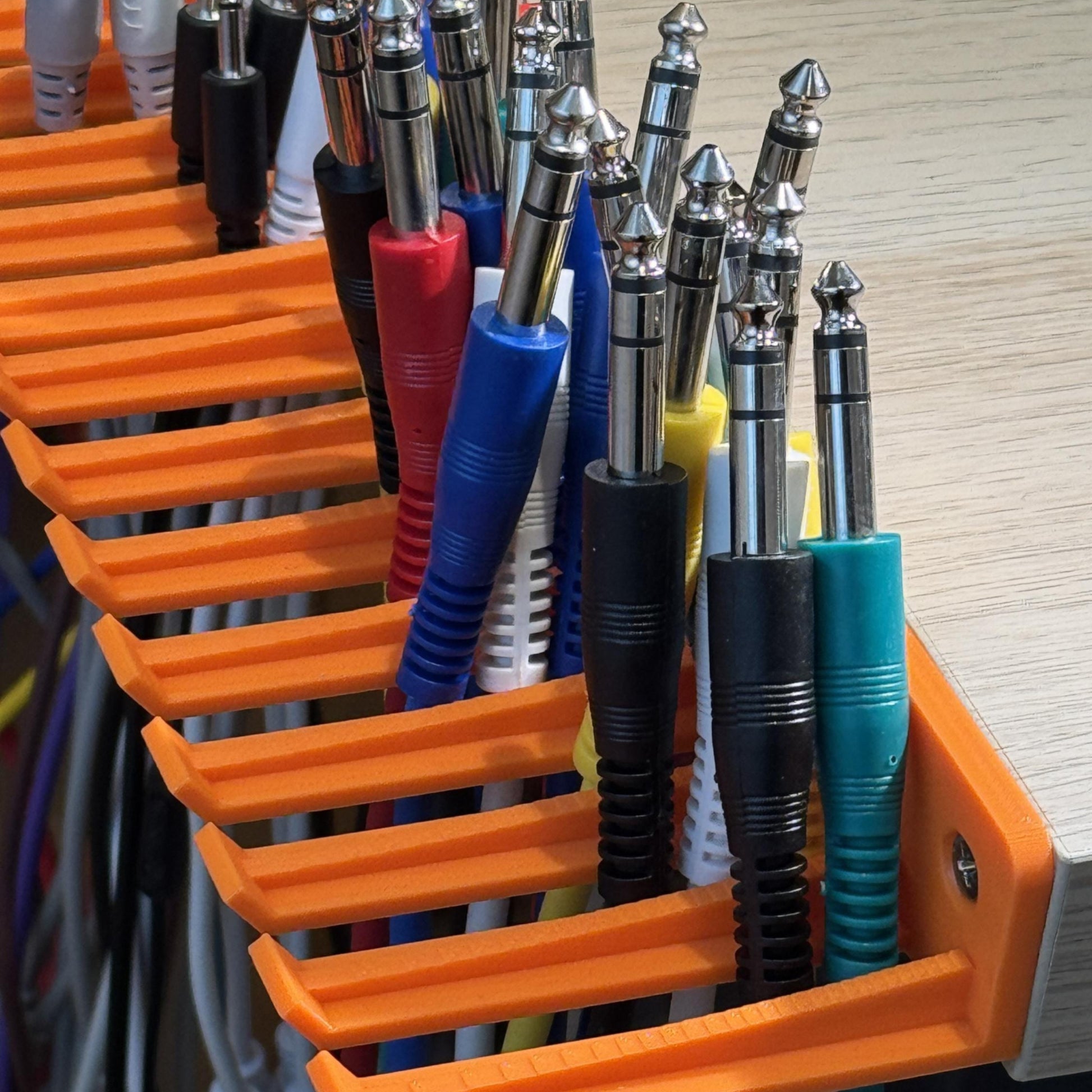 3D printed Guitar Pedal Patch Cable Holder by Cold Earth Industries, shown with cables, in orange, screwed to the side of a desk – custom studio and live performance gear.