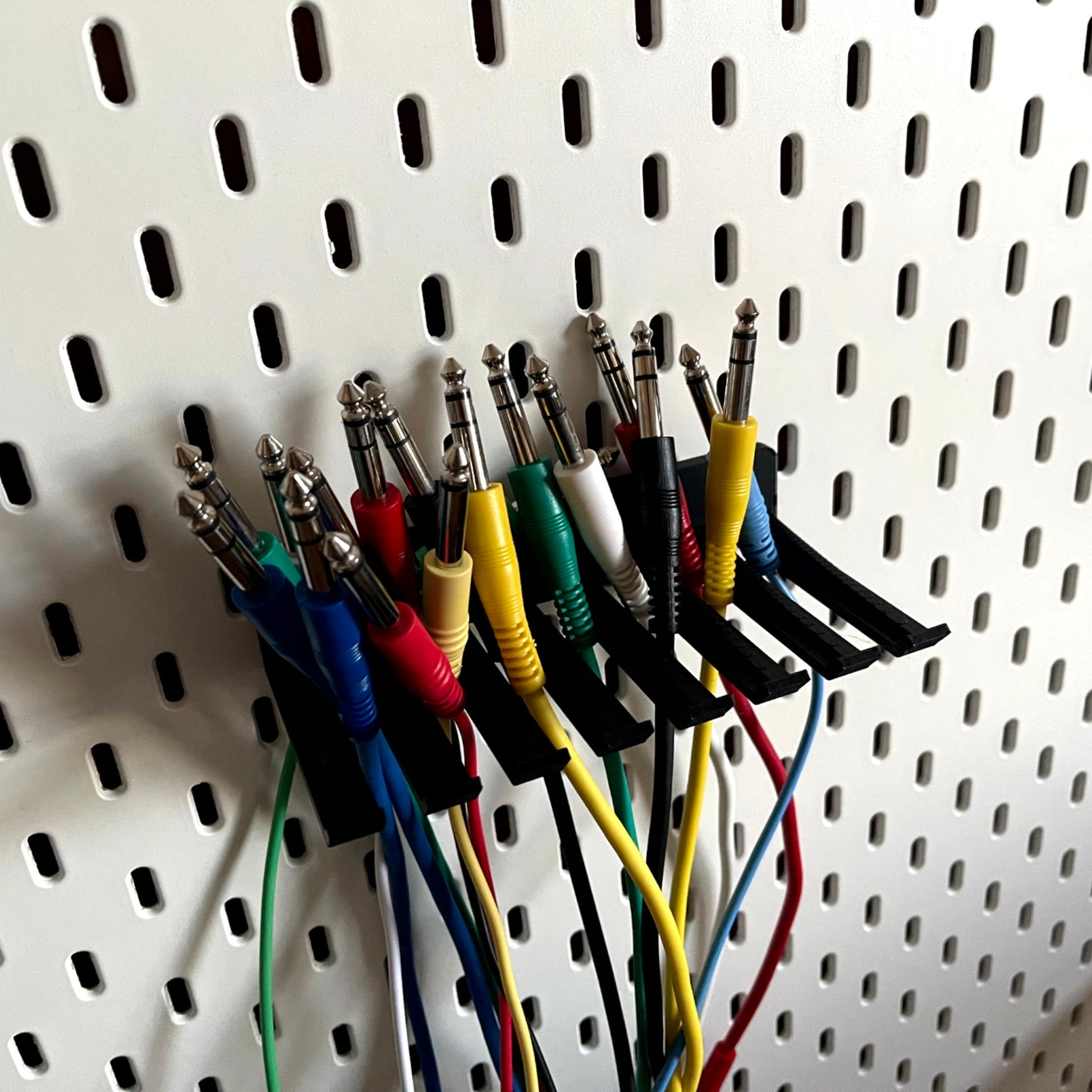 3D printed Guitar Pedal Patch Cable Holder for Ikea Skadis by Cold Earth Industries, holding guitar cables, shown in black on a white Ikea Skadis Pegboard – custom studio and live performance gear.