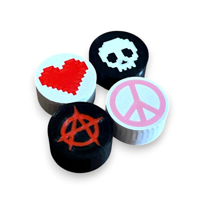 3D printed guitar pedal footswitch toppers with various designs including a heart, skull, anarchy symbol, and peace symbol, printed in black with colorful icons.